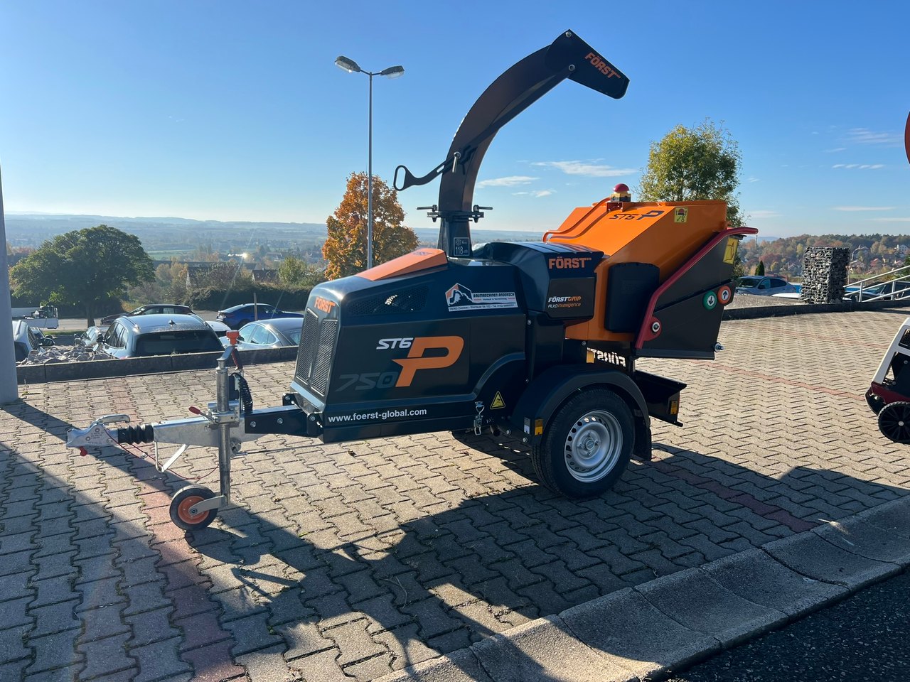 Ein orange-schwarzer Forst ST6P Holzhäcksler auf einem Anhänger, unter blauem Himmel.