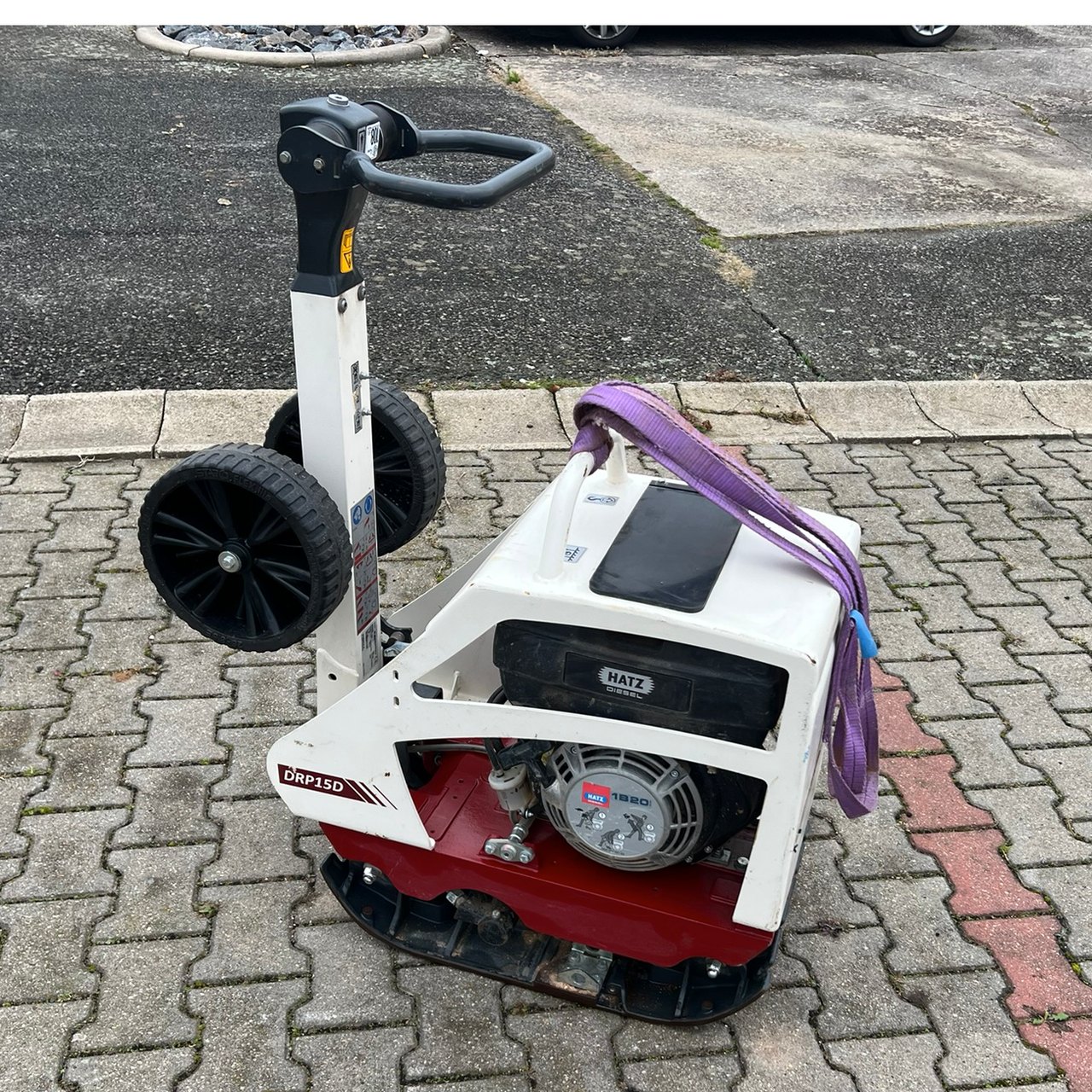 Eine weiße und rote Rüttelplatte mit Griff, Rädern und lila Spanngurt auf grauen Pflastersteinen.