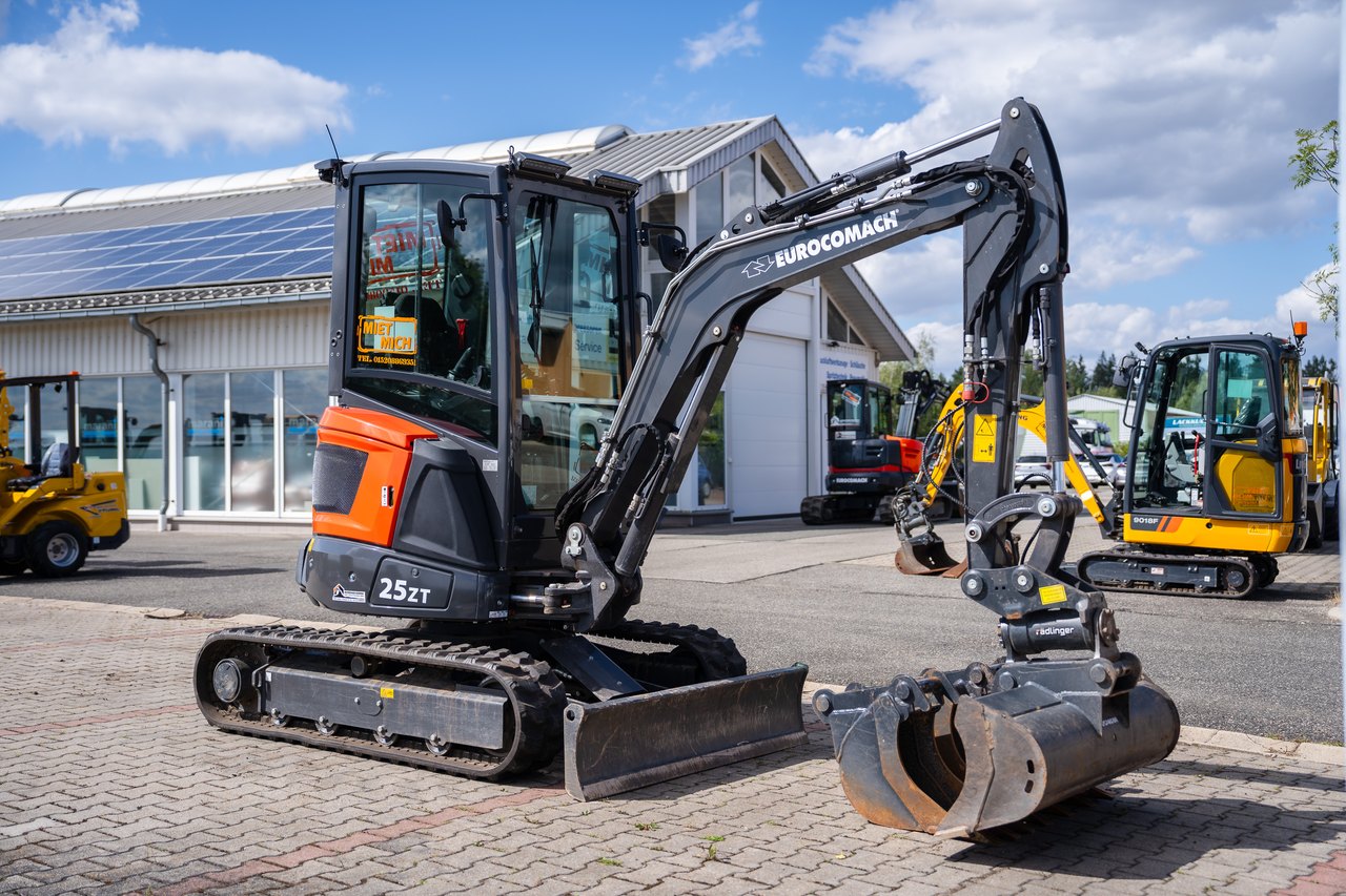 Schwarzer Euromach 25ET Minibagger mit Greifer auf asphaltiertem Parkplatz vor einem Gebäude.