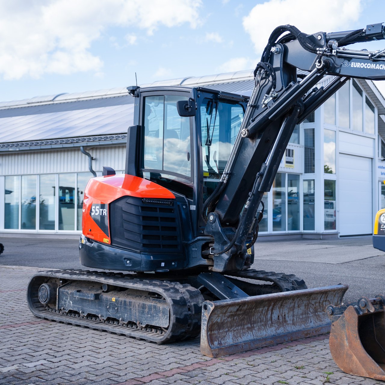 Oranger und schwarzer Kubota 55TR Minibagger auf einem gepflasterten Hof vor einem Gebäude.