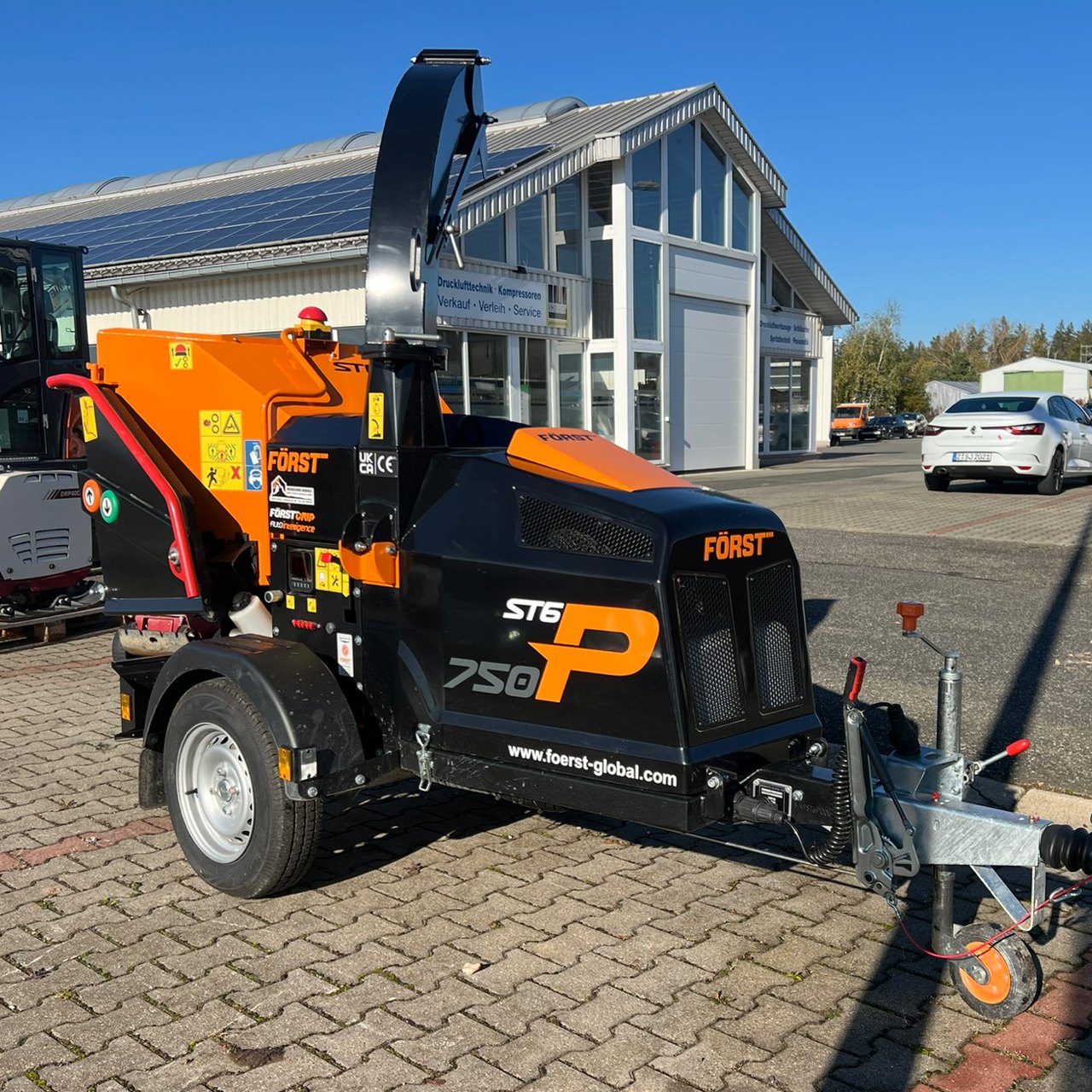Ein schwarzer und oranger Först ST6 750 Holzhacker auf einem Anhänger, geparkt vor einem Gebäude mit Solarpaneelen.