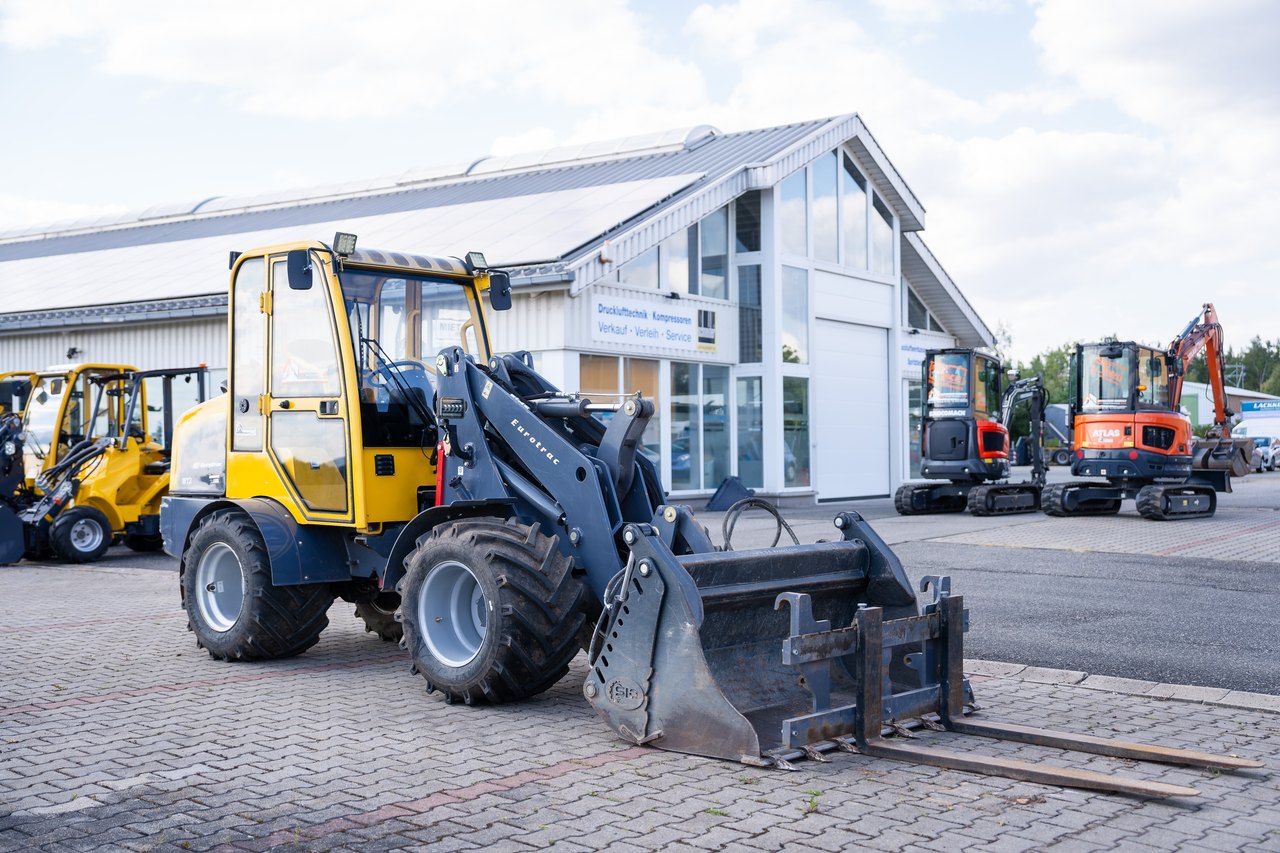 Gelber Radlader mit Gabelaufsatz vor einem Gebäude, weitere Baumaschinen im Hintergrund.