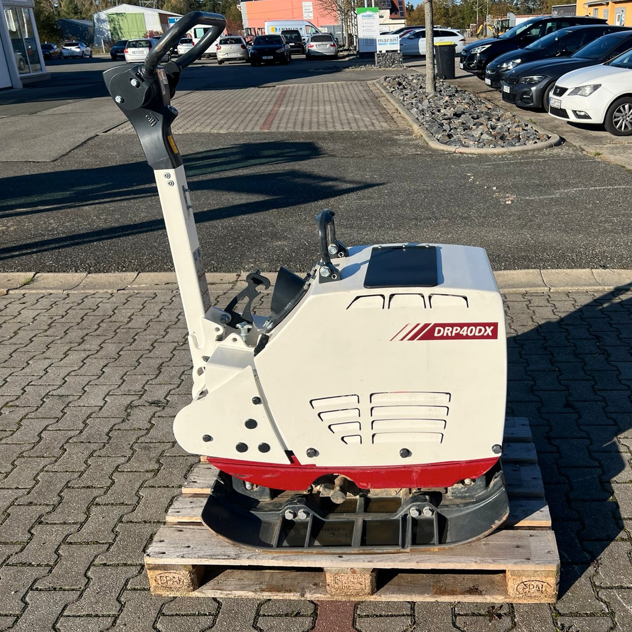 Weiße und rote Rüttelplatte DRP40DX auf einer Holzpalette auf einem gepflasterten Parkplatz.